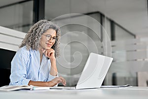 Happy mature professional business woman looking at laptop working in office.
