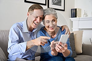 Happy older mature couple enjoying using cell phone sitting on couch at home.