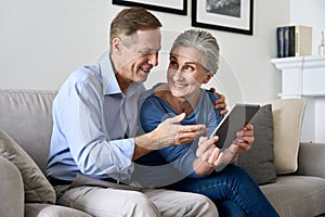 Happy older mature couple using digital tablet sitting on sofa at home.