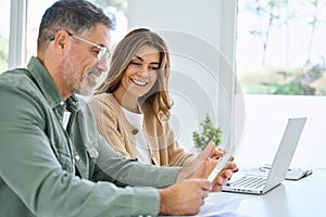 Happy older mature couple using laptop counting taxes with receipts at home.