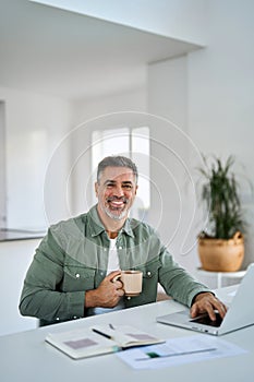 Happy older mature man drinking coffee using laptop at home table. Portrait.