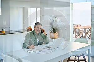 Happy older mature man talking on phone using laptop at home.