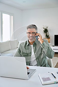 Happy older mature man talking on phone using laptop at home. Vertical.