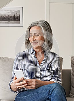 Happy older mature woman enjoying using cell phone sitting on couch at home.