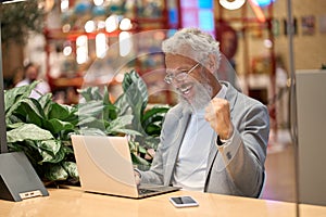 Happy older business man using laptop celebrating good results winning online.