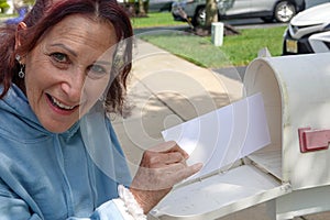 Happy older senior citizen woman smiles as she removes a blank white envelope from a white mailbox