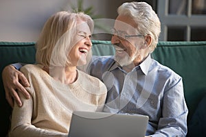 Happy older spouses couple laughing embracing using laptop at home