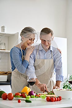 Happy older vegan couple preparing healthy vegetable salad at home in kitchen.