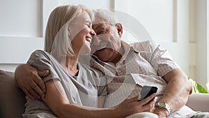 Happy older wife and husband cuddling, using phone together