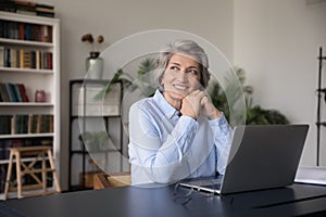 Happy older woman sit at workplace desk looking into distance