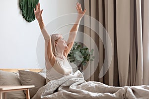 Happy older woman sitting in bed, stretching hands after awakening