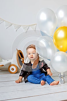 A happy one-year-old boy in a helmet and a pilot`s jacket sits near a wooden plane and balloons. Portrait in the studio of a chil