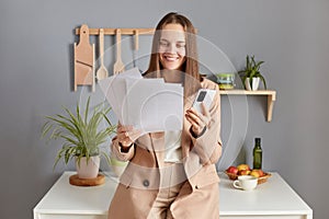 Happy optimistic woman freelancer with brown hair wearing beige jacket standing in kitchen interior using mobile phone checking