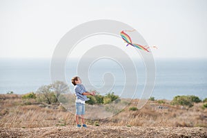 Happy outdoor leisure games by small active caucasian boy with flying kite in air with sea horizon and blue sky in summer