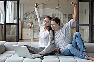 Happy overjoyed couple celebrating online betting bid lottery win sitting on couch at home with raised hands
