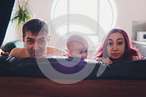 Happy parents with adorable little baby girl sitting on the couch at home, enjoying parenthood, young happy family, new