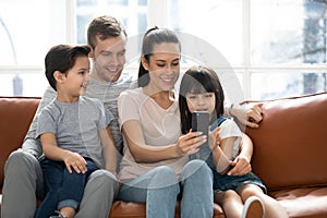 Happy parents with cute kids sit on sofa using phone