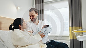 Happy parents holding newborn baby boy in hospital ward. Smiling mother sitting in hospital bed with husband near and
