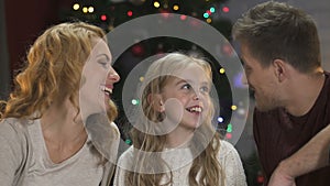 Happy parents kissing their adorable daughter on cheeks sitting under x-mas tree