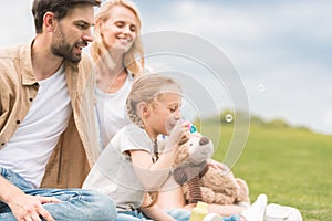 happy parents looking at cute little daughter with teddy bear blowing soap bubbles