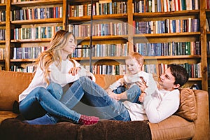 Happy parents playing with small child at Christmas time at home in living room on sofa near large cabinet with books and