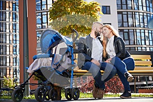 Happy parents sitting on bench with stroller in autumn.