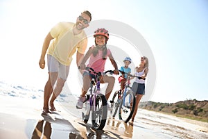 Happy parents teaching children to ride bicycles on beach near sea