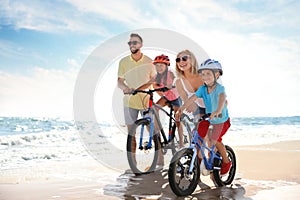 Happy parents teaching children to ride bicycles on sandy beach near sea