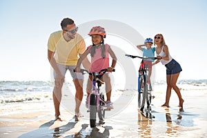 Happy parents teaching children to ride bicycles on sandy beach near sea