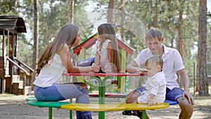 Happy parents with their little children are resting on playground in park with high spruce in summer day