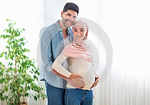 Happy parents-to-be. Happy arab couple waiting for baby, embracing and smiling to camera, standing near window