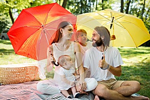 Happy parents with two kids have a rest on the lawn under the bright red and yellow umbrellas covering them from the sun