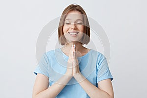 Happy peaceful beautiful young woman in blue t shirt keep hands folded in praying position and eyes closed isolated over white