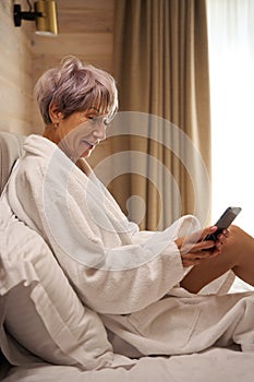 Happy pensioner in bathrobe sits in bed with mobile phone