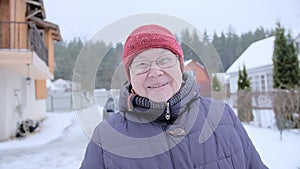 Happy pensioner at the dacha. Grandmother in glasses, a red knitted hat and a blue jacket smiles at the camera