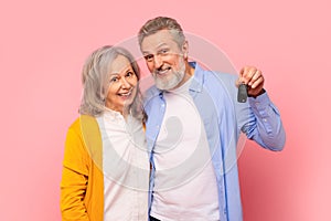 Happy pensioners couple excitedly showing automobile key on pink backdrop