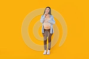 Happy pensive pregnant woman touching belly and chin, looking up and thinking, standing on yellow studio background