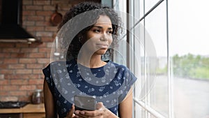Happy pensive young African American woman with smartphone looking away