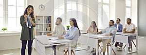 Business coach teaching group of adult students who are sitting at desks in classroom