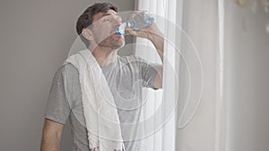 Happy perspiring middle aged man with towel on shoulder drinking water, wiping mouth with hand, and looking at camera