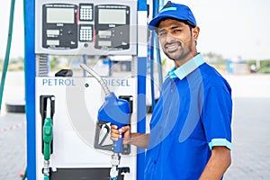 Happy petrol pump worker standing by holding fuel nozzle while looking camera at gas filling station - concept of