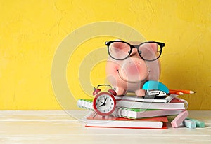 Happy piggy bank with glasses, and school accessories on white wooden table against color background, space for text