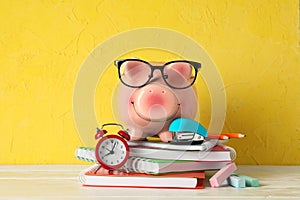 Happy piggy bank with glasses, and school accessories on white wooden table against color background, space for text