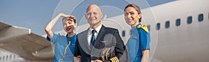 Happy pilot and two attractive stewardesses standing together in front of an airplane and smiling after landing