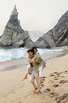 Happy playful young couple having fun on beach, man giving piggyback ride to his girlfriend, enjoying date on coastline