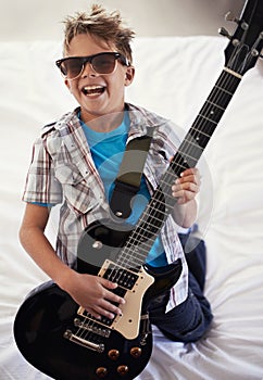 Happy, playing and boy kid with guitar on bed for music lesson with sunglasses at modern home. Fun, rockstar and young