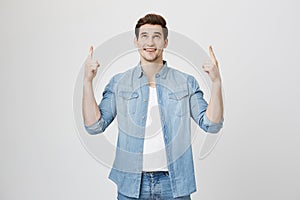 Happy pleased Caucasian male with stubble points with forefingers upwards dressed in denim clothes, smiles joyfully photo