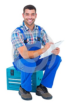 Happy plumber writing on clipboard while sitting on toolbox