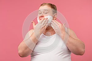 Happy plump man with shaving foam on his face and razor isolated on pink
