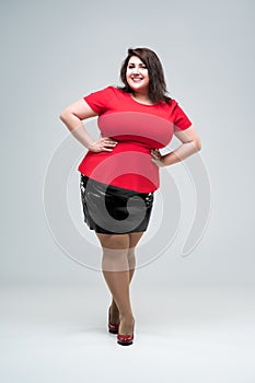 Happy plus size fashion model in red blouse and black skirt, fat woman on gray background, body positive concept